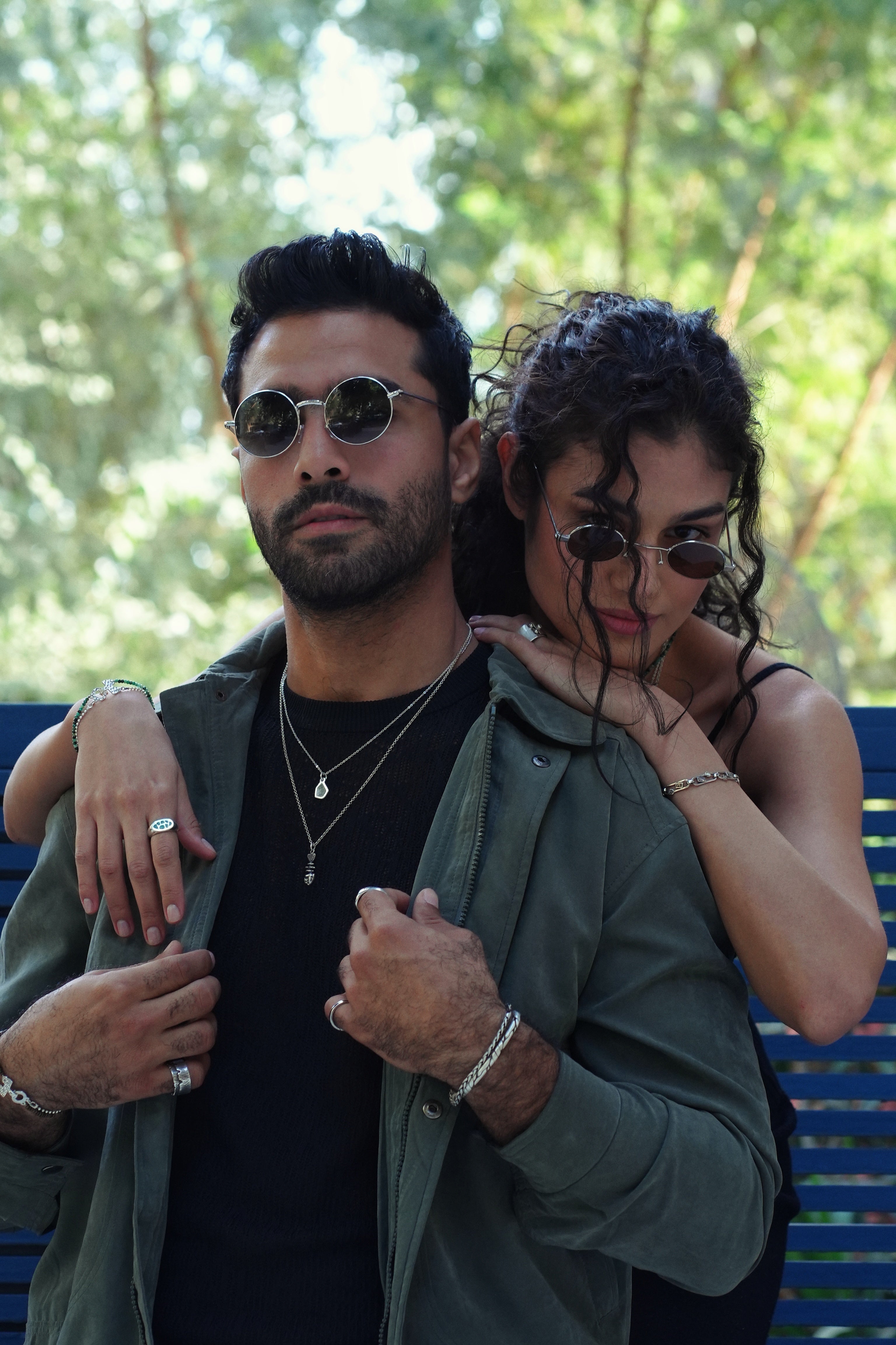 Man and woman posing together for silver jewlery on a bench with trees in the background